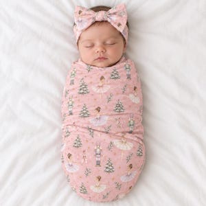 May include: A swaddle blanket and matching headband set in a soft pink fabric featuring a repeating pattern of nutcrackers, ballerinas, evergreen trees, and gold stars. The fabric is wrapped around a sleeping infant, and the bow headband is worn.