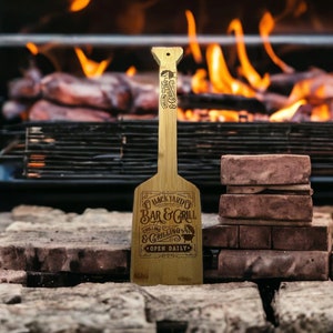 May include: A wooden spatula with the text "Backyard Co Bar & Grill" is displayed in front of a grill with flames. The spatula has a long handle and a wide, flat head. The text "Open Daily" is also visible.
