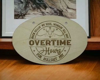 Puede incluir: Un letrero de madera ovalado de color marrón claro con el texto "BEEN SELLIN' MY SOUL, WORKIN' ALL DAY * OVERTIME * Hours FOR BULLSHIT PAY" en una fuente marrón. Un cráneo estilizado con cuernos está por encima de la palabra "OVERTIME".