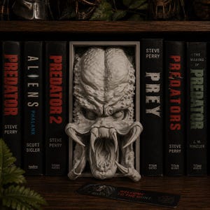 May include: A light grey, rectangular bookend featuring a sculpted, detailed Predator creature head with an open mouth and prominent mandibles. The bookend is positioned between several black books with red and white text on their spines.