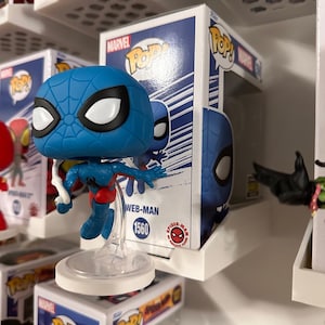 May include: A blue Web-Man Funko Pop! vinyl figure with black eyes and a red and blue suit. The figure is displayed on a white shelf, with the box behind it. The box has the Marvel and Pop! logos, and the number 1560.