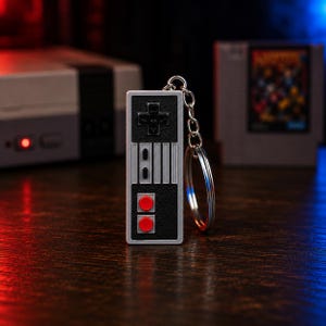 May include: A rectangular keychain designed to resemble a retro video game controller. The device features a black textured surface with a silver border, a black directional pad, two small black buttons, and two prominent red circular buttons.