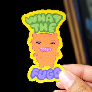 May include: A sticker featuring a cartoon monster with orange fur, a purple mouth, and the words "WHAT THE FUCC" in green and purple. The sticker has a yellow border and is held by a hand.