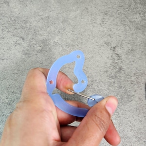 May include: A light blue earring holder with a curved design and small star-shaped cutouts. A silver earring post is inserted into a small, round earring. The holder is held in a hand against a gray background.