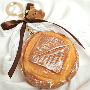 May include: A round, bread-shaped keychain with a leaf design on top. It features a brown ribbon, white lace, and a small teddy bear charm. The keychain has a gold-colored ring and clasp.