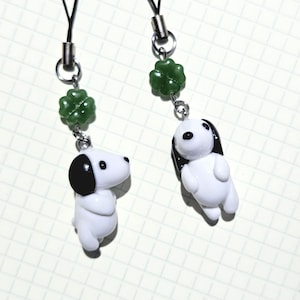 May include: Two white and black dog-shaped charms with green clover beads, attached to black cords. The charms have black ears and eyes. The clover beads are a dark green color.