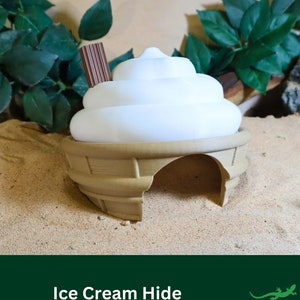May include: A white ceramic ice cream cone shaped hide for small reptiles. The hide has a brown, textured base and a white, smooth top. The hide is sitting on a bed of sand.