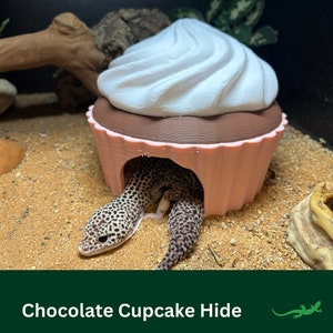 May include: A pink and brown 3D printed cupcake hide for small reptiles, with a white frosting top. A leopard gecko is partially inside the hide.