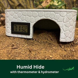 May include: A white plastic reptile hide with a textured surface. The hide has a built-in digital thermometer and hygrometer that reads 71 degrees and 21%. The hide is sitting on a brown dirt surface.