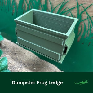 May include: A green plastic frog ledge shaped like a small dumpster. The ledge is designed to be mounted on a wall or terrarium.