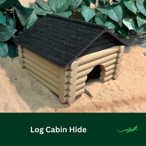May include: A small log cabin hide made of brown wood with a black roof. The hide is sitting on a sandy surface.