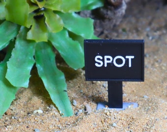Può includere: Un cartello nero con testo bianco che dice "SPOT" è piantato nella sabbia accanto a una pianta verde.