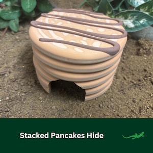 May include: A reptile hide shaped like a stack of pancakes, with a brown drizzle design. The hide is tan and brown, with a circular entrance. The text "Stacked Pancakes Hide" is at the bottom. The hide is designed to provide shelter.