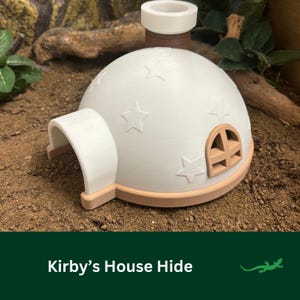 May include: A white dome-shaped hide with a brown base and a star pattern. The hide has a small window and a tunnel entrance. The hide is on a brown dirt surface. Kirby's House Hide.