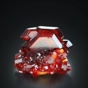 Può includere: Un gruppo di cristalli di vanadinita rossi con un grande cristallo ben formato al centro. I cristalli sono molto riflettenti e hanno una forma geometrica appuntita.