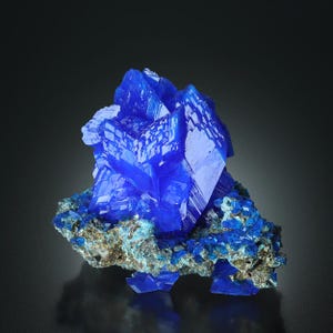 May include: A vibrant blue mineral specimen with crystalline structures. The mineral has a deep, saturated blue color, with sharp, well-defined crystal formations. The base rock is a mix of brown and blue-green hues, providing a contrasting backdrop.