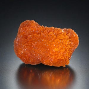 May include: A vibrant orange mineral specimen with a crystalline structure. The mineral has a rough, textured surface and reflects light, showcasing its intricate formations. The color is a deep, saturated orange, with a slight translucence.
