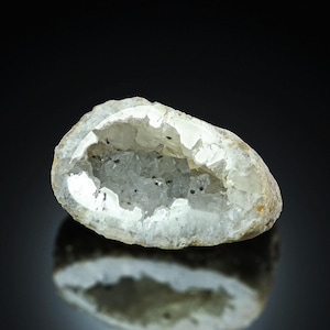 Könnte beinhalten: Eine kleine, oval geformte Geode mit einer rauen, hellen Außenseite und einem hohlen Zentrum, das mit funkelnden, durchscheinenden weißen Quarzkristallen gefüllt ist. Der innere Hohlraum zeigt gezackte Kristallformationen mit dunklen Einschlüssen.