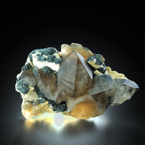 May include: A cluster of smoky quartz crystals with a dark, translucent appearance. The crystals are set against a backdrop of green and white mineral formations, with a touch of yellow. The overall composition is a natural, earthy display.