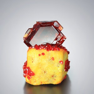 Peut inclure: Un groupe de cristaux rouge vif sur un spécimen minéral jaune. Les cristaux sont translucides et ont un éclat vitreux.