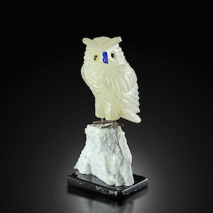May include: A white carved owl figurine with blue and yellow eyes, perched on a white rock base. The owl is made of alabaster and is a decorative piece.