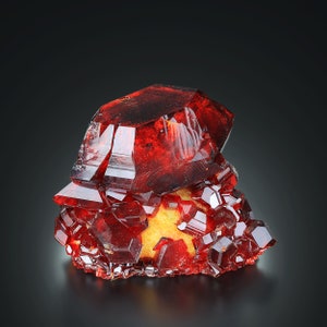 May include: A cluster of deep red, translucent crystals with a sharp, geometric shape. The crystals are arranged in a pyramid-like formation, with the largest crystal at the top.