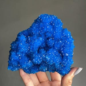 May include: A vibrant, crystalline cluster of deep blue crystals. The mineral formation has a rough, textured surface with numerous small, sparkling facets. The overall shape is irregular and organic, with a rich, saturated color. The crystals are held in a hand.