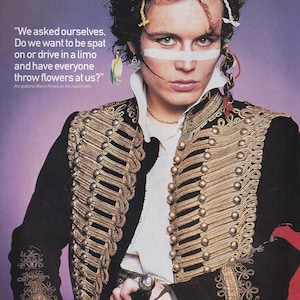 Op de afbeelding: Een portret van een persoon in een zwarte jas met gouden details, een wit overhemd en een witte streep over het gezicht. De jas heeft gouden vlechtwerk en knoopdetails. De afbeelding bevat de quote: "We vroegen ons af, Willen we bespuugd worden of in een limousine rijden en dat iedereen bloemen naar ons gooit?"