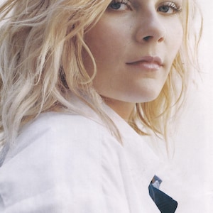May include: A portrait of a person with blonde, wavy hair. The person is wearing a white collared shirt. The image is a close-up, focusing on the person's face and upper body.
