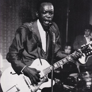 Könnte beinhalten: Schwarz-Weiß-Foto eines Musikers, der auf der Bühne eine weiße E-Gitarre spielt und in ein Mikrofon singt. Der Text im Bild lautet: "Bringing the boogie to England, 1964. By now a whole generation of musicians had learned his songs."