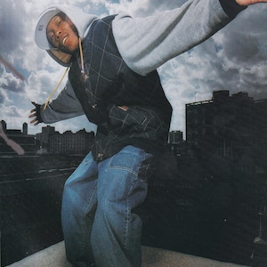 Può includere: Un giovane uomo con una giacca con cappuccio, jeans e scarpe da ginnastica posa con il braccio teso sullo sfondo di un cielo nuvoloso e di edifici cittadini. Il testo "Top of the world, mal Dizzee Rascal photographed exclusively for MOJO, 5th and Sunset Studios, London N1, September 14, 2004." è stampato nella parte inferiore dell'immagine.