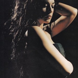 May include: A portrait of a person with long, dark, wavy hair. The person is wearing a dark gray, button-down top. The image is set against a dark background, with the person's face and arm illuminated.