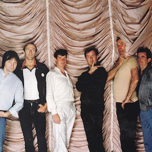 May include: Seven men stand in front of a draped backdrop. They wear casual clothing, including shirts, jackets, and trousers. The color scheme is subdued, featuring white, black, blue, and beige. The image has a vintage aesthetic.