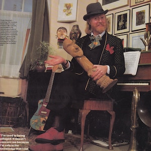 May include: A person wearing a dark bowler hat, a black patterned blazer with gold trim, a dark vest, and a patterned bow tie. They are holding a light brown carved wooden sculpture of a face and torso while sitting on a wooden stool.