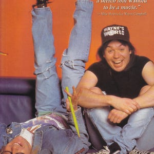 May include: A promotional image featuring two men from the movie "Wayne's World." One man is upside down, wearing a denim jacket and jeans. The other man is wearing a "Wayne's World" hat and black t-shirt. The quote "This was always a sketch that wanted to be a movie" is visible.