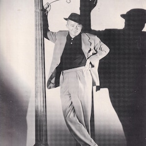 May include: A black and white photograph featuring a person wearing a hat, a dark collared shirt, a light-colored blazer, and trousers, leaning against a street sign post labeled SUNSET BLVD. The text King Billy Wilder: A wicked, wicked wit is visible.
