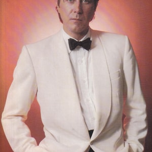 May include: A man in a white tuxedo jacket with a black bow tie. He is standing against a red background. The text "Re-releases" is visible in the top left corner of the image.