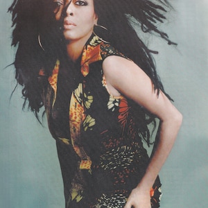 Può includere: Una donna con capelli lunghi e scuri indossa una canotta colorata con motivi e un fondo a frange. La canotta ha uno sfondo nero con accenti gialli, arancioni e rossi.