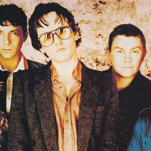 May include: A group of five people pose for a portrait. The central figure wears glasses and a tweed jacket over a button-down shirt. Others are in various shirts and jackets. The background is a mottled, neutral tone.