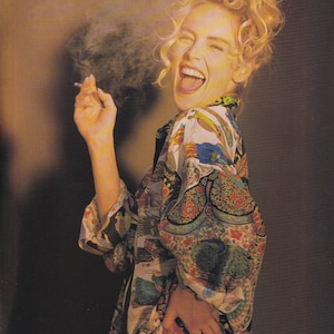 Może przedstawiać: Kobieta o blond, kręconych włosach nosi kolorową, wzorzystą koszulę. Uśmiecha się i trzyma papierosa w prawej ręce. Z papierosa unosi się dym.