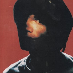 May include: A close-up of a person's face with dark hair and a dark shirt. The background is a solid red colour. The text "Jonny Greenwood: guitarist in the shadows." is at the bottom of the image.