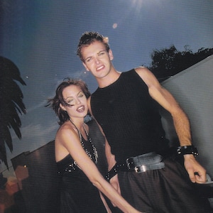 Könnte beinhalten: Ein Paar posiert vor einer weißen Wand für ein Foto. Der Mann trägt ein schwarzes Tanktop und eine schwarze Hose mit einem schwarzen Gürtel. Die Frau trägt ein schwarzes Kleid mit einer silbernen Halskette. Der Mann hat kurze braune Haare und die Frau hat lange braune Haare.