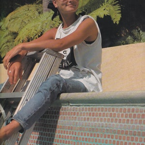 Könnte beinhalten: Eine Person mit einem weißen Tanktop mit Grafikdesign, blauen Jeans und Sonnenbrille sitzt auf einem Sims neben einem Pool. Ein schwarzes Bandana bedeckt den Kopf der Person. Eine Metallleiter befindet sich neben der Person.