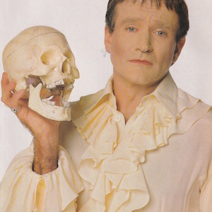 May include: A man in a light yellow ruffled shirt holds a human skull. The skull is off-white and has visible teeth. The man has short brown hair and is looking at the camera.