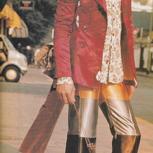 May include: A person in a vintage outfit, wearing a red jacket, floral shirt, and patchwork trousers. The trousers have silver, black, and gold panels. White shoes complete the look. The image is from a magazine.