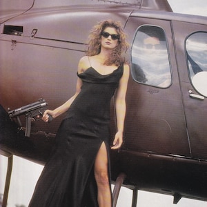 Może przedstawiać: Kobieta w czarnej sukience i okularach przeciwsłonecznych stoi obok brązowego helikoptera, trzymając pistolet maszynowy. Sukienka ma wysoki rozporek. Helikopter stoi na trawniku.