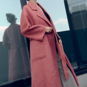 May include: A long, dusty rose-colored coat with a wide lapel and large pockets. The coat is belted at the waist and has long sleeves. The coat is made of a soft, textured material. The coat is part of a fashion collection.