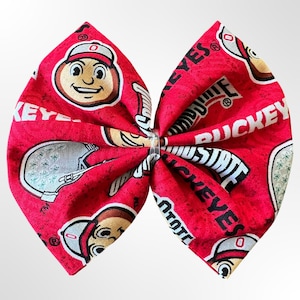 May include: A large, red fabric bow featuring a repeating pattern of the Ohio State Buckeyes mascot, helmet, and text. The bow is tied in the center with a clear plastic fastener. The design includes white, black, and tan accents.