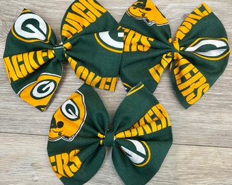 Puede incluir: Tres lazos para el pelo de tela verde y dorada con el logotipo de los Green Bay Packers y la palabra "Packers" impresa en ellos.