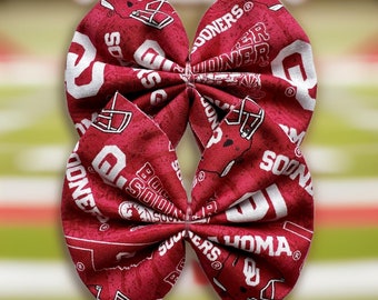 May include: A red and white fabric bow with a University of Oklahoma logo and the words "Sooners" and "Oklahoma".